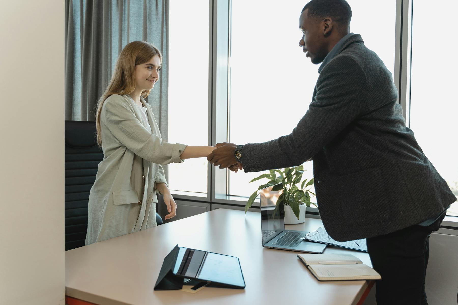 Two business professionals shaking hands across a desk in a bright office